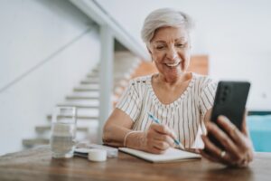 Older woman viewing mobile phone while writing paper notes with a pen